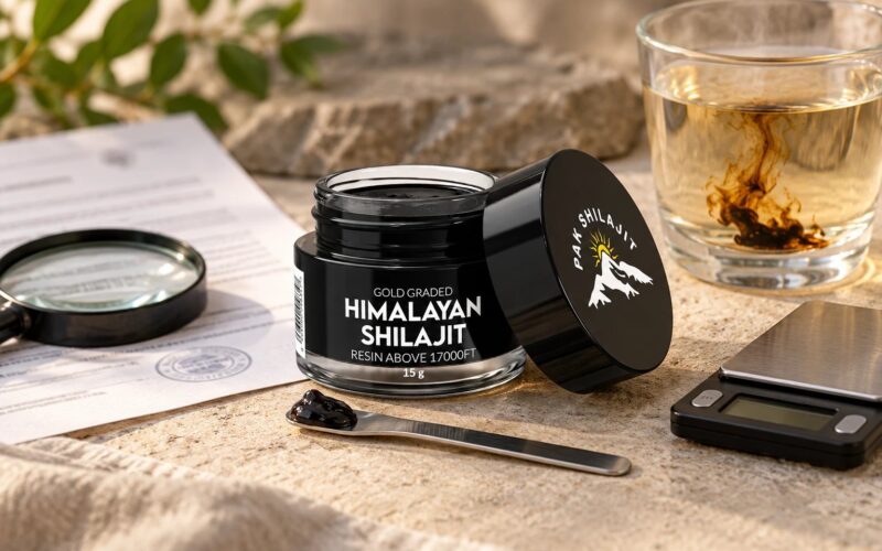 Pak Shilajit resin jar on a stone worktop beside a printed lab report under a magnifying glass, the steel spatula holding a pea-sized piece of resin, a glass of warm water with resin dissolving, and a pocket scale