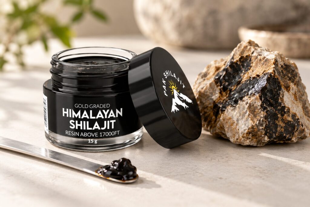 Open Pak Shilajit jar with a steel spatula holding a piece of dark resin, beside a raw piece of Himalayan rock veined with shilajit