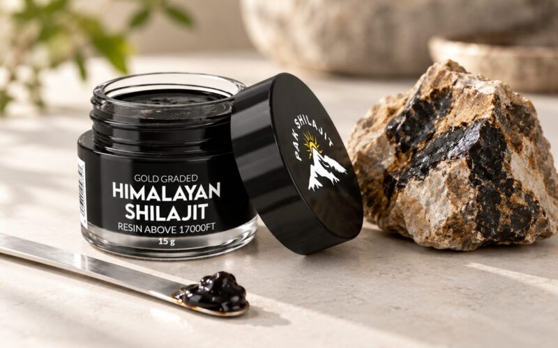 Open Pak Shilajit jar with a steel spatula holding a piece of dark resin, beside a raw piece of Himalayan rock veined with shilajit