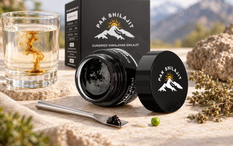 Pak Shilajit resin jar with the steel spatula holding a pea-sized serving of resin, a grain of rice and a pea beside it for scale, and a glass of water