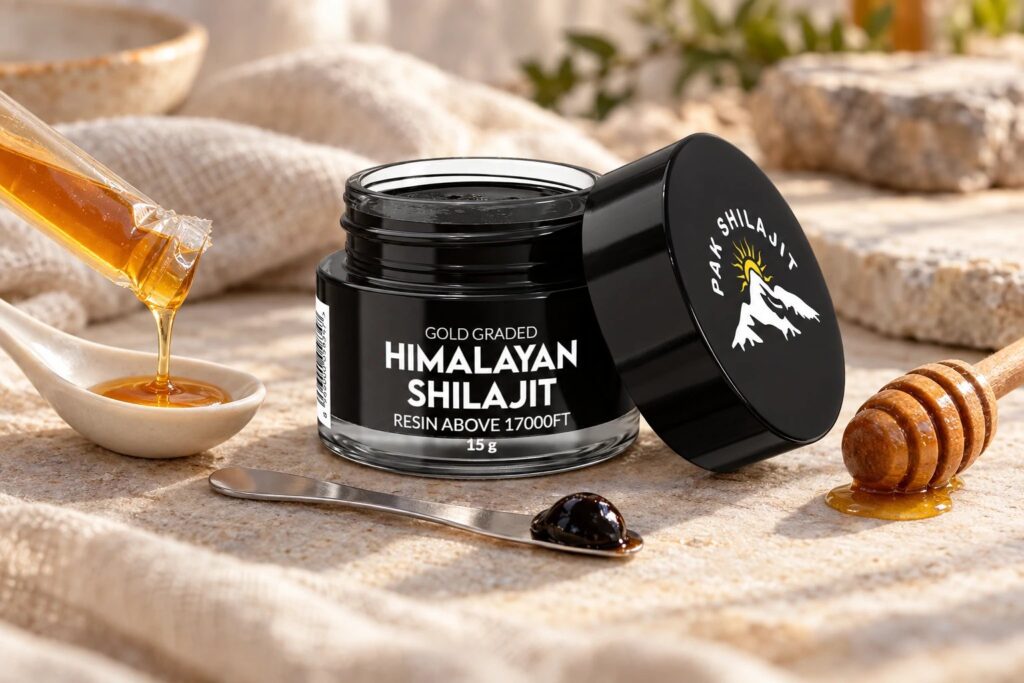 Pak Shilajit resin jar open on linen beside a torn honey stick dripping into a ceramic spoon, a wooden honey dipper, and the steel spatula holding a pea-sized piece of resin
