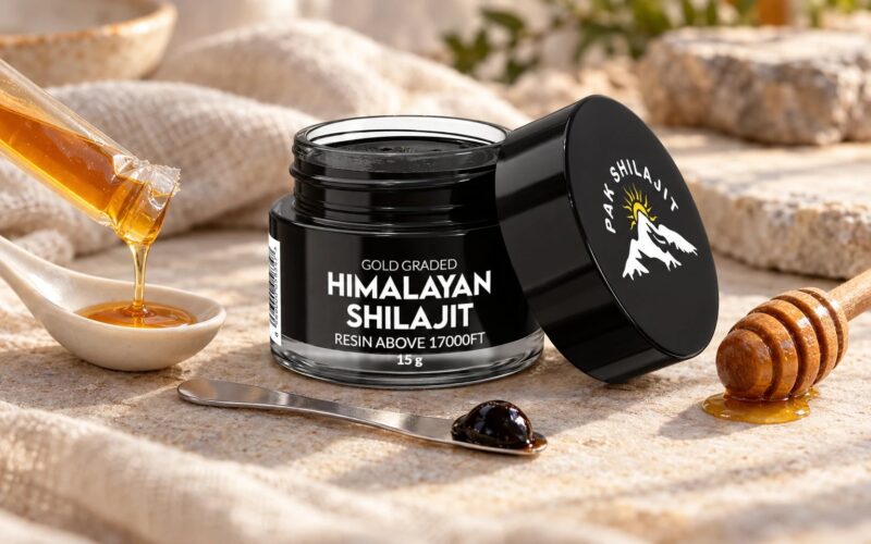 Pak Shilajit resin jar open on linen beside a torn honey stick dripping into a ceramic spoon, a wooden honey dipper, and the steel spatula holding a pea-sized piece of resin