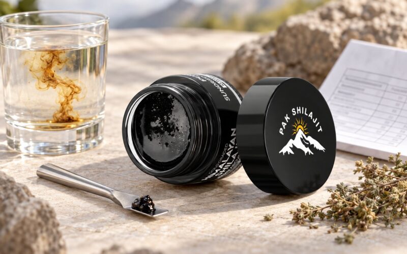 Pak Shilajit resin jar beside a glass of water, a measuring spoon holding a rice-grain serving, and a folded lab report