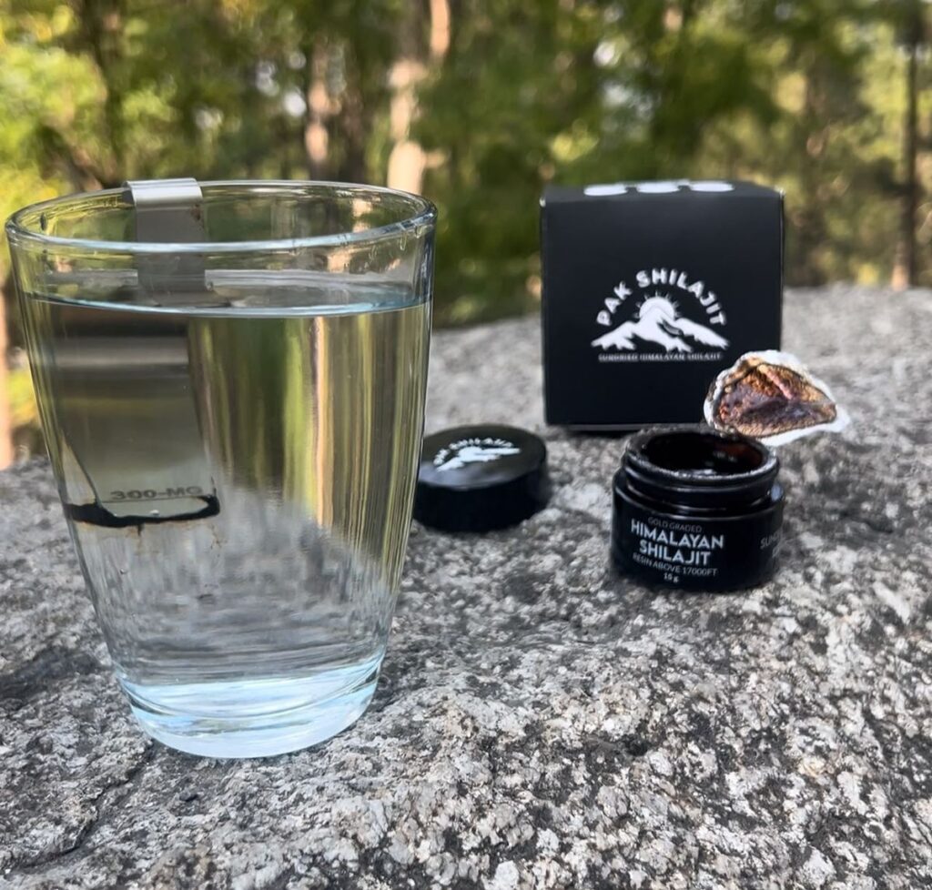 A piece of Pak Shilajit resin suspended in a glass of clear water beside the jar and box, before dissolving begins