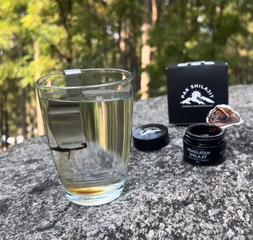 Shilajit resin dissolving in warm water, a dark ribbon running down and amber colour gathering at the base of the glass